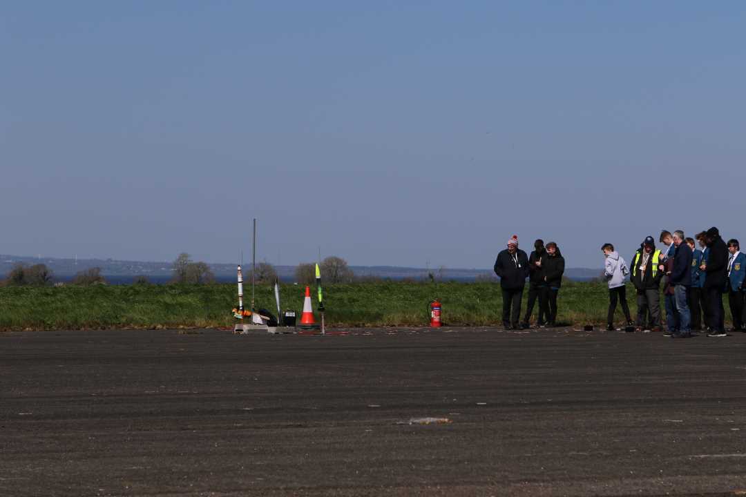 3 model rockets sitting on pads with a small crowd standing at a distance to the right